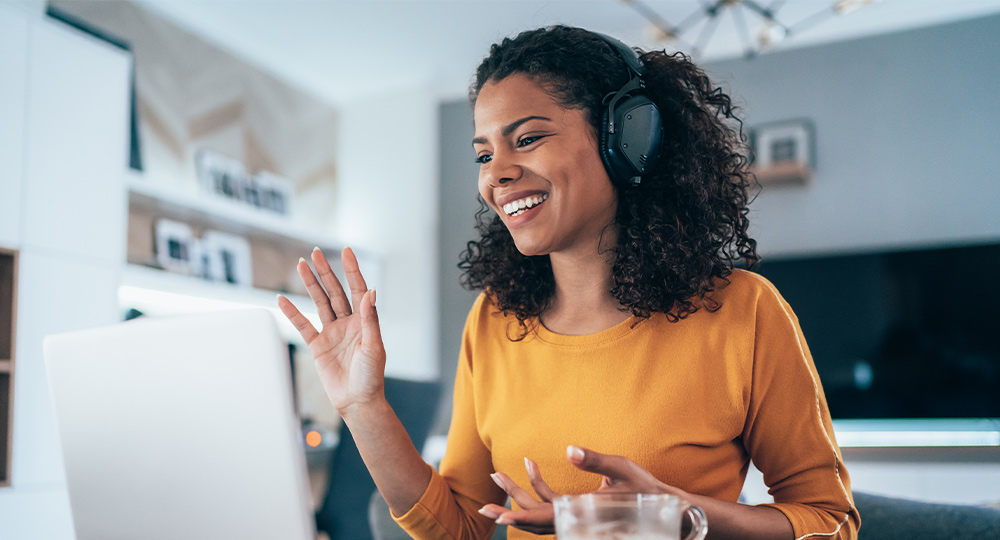 Woman with headphones on smiling and waving at someone on the computer in a teleconference.