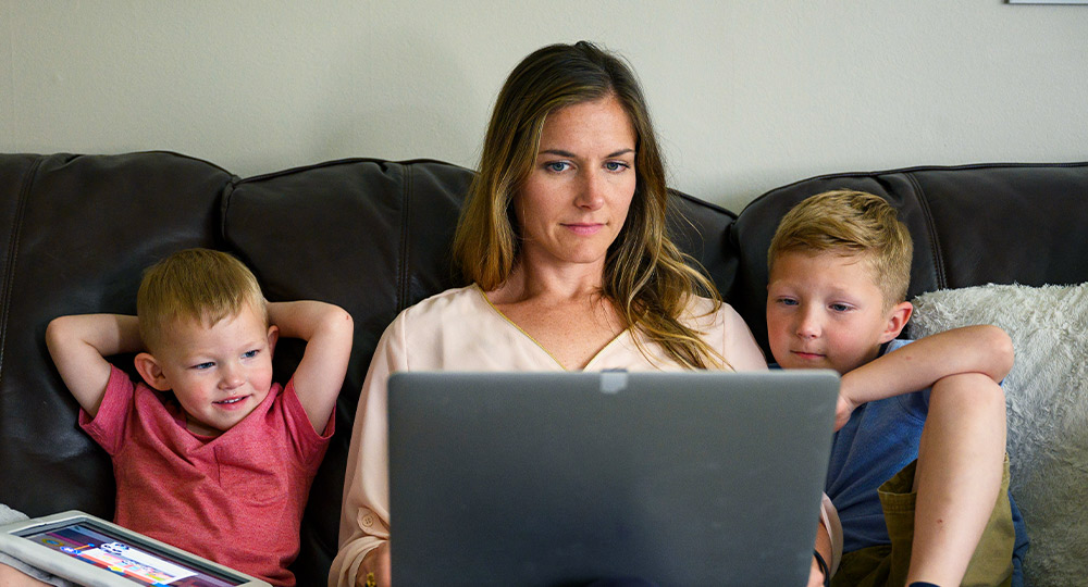 Woman sitting on the couch with her two kids while using her computer.