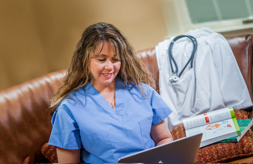 Industry image of a woman in a health program with a laptop in scrubs with textbooks and labcoat